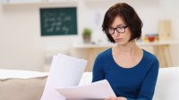 A female teacher in a blue shirt and glasses is sitting in a classroom alone, looking at papers.