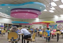 Young students are working at tables in a large multipurpose room with large colorful-shaped decorations hanging from the ceiling. 